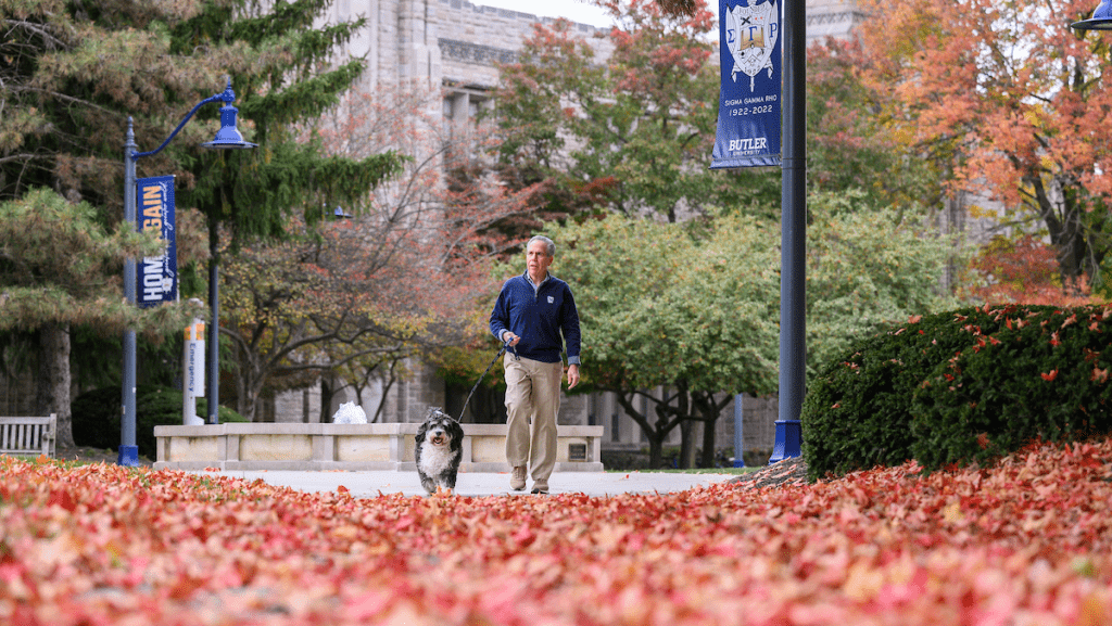 President Danko in navy sweater and khaki slacks walking through fall leaves with black and white dog on leash