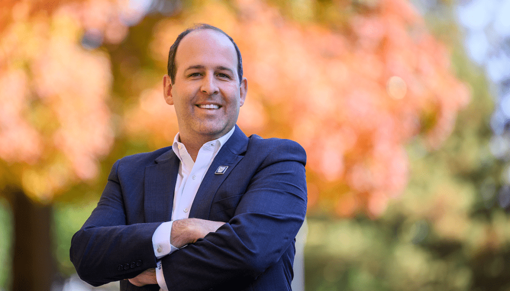 male with arms crossed, wearing blue jacket and white shirt, outside in front of gold leaves on tree
