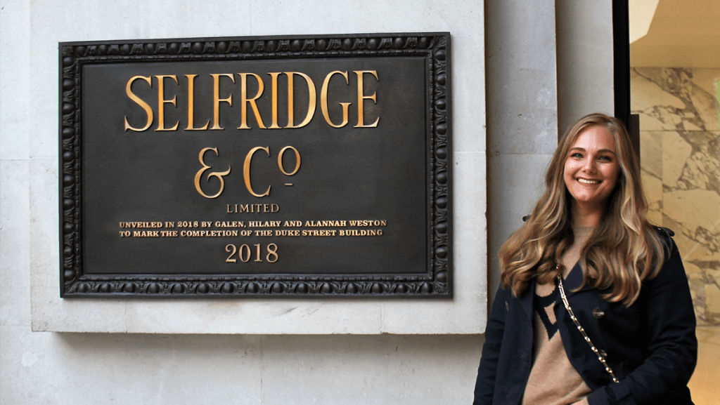 large sign on concrete wall reading Selfridge & Co Limited. Blonde woman next to sign in dark jacket