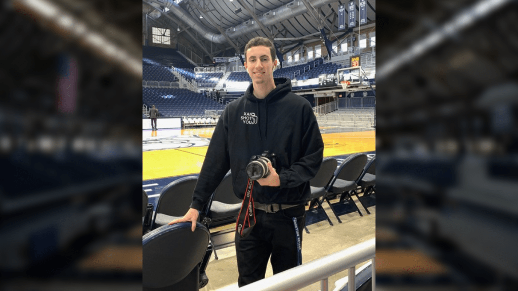 Male in dark sweatshirt with words Xan Shot You, holding camera, in HInkle Fieldhouse