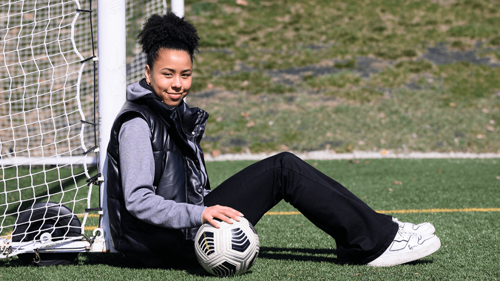 female in dark pants and vest sitting in front of soccer net, with soccer ball