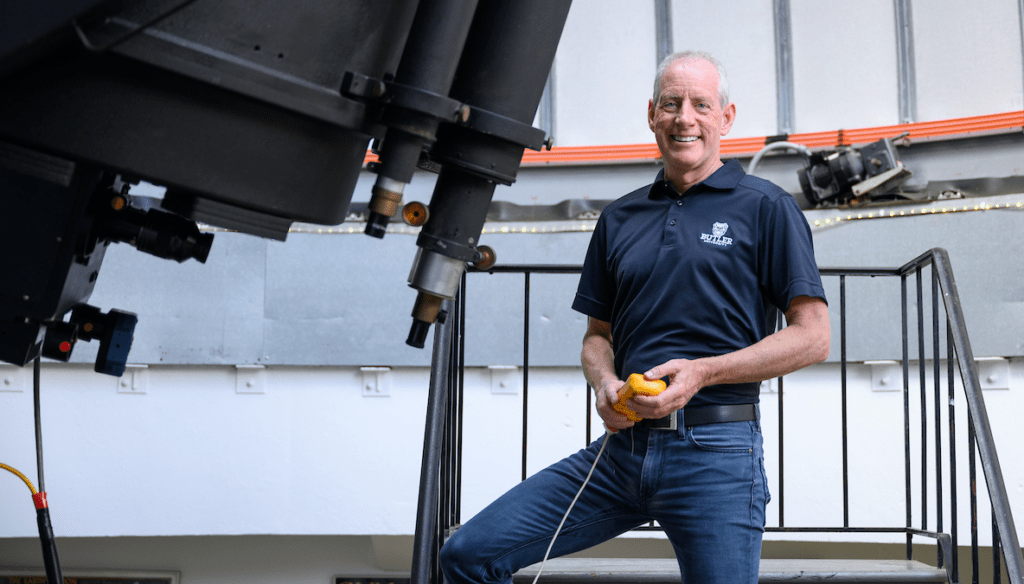 man in blue jeans and blue shirt with Butler logo standing at large telescope.