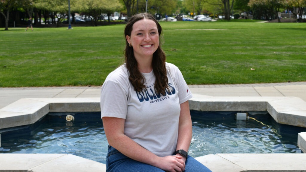 Butler student, Sarah Mangan, wearing a white Butler t-shirt and jeans, posing for a picture sitting at Star Fountain on Butler's campus.