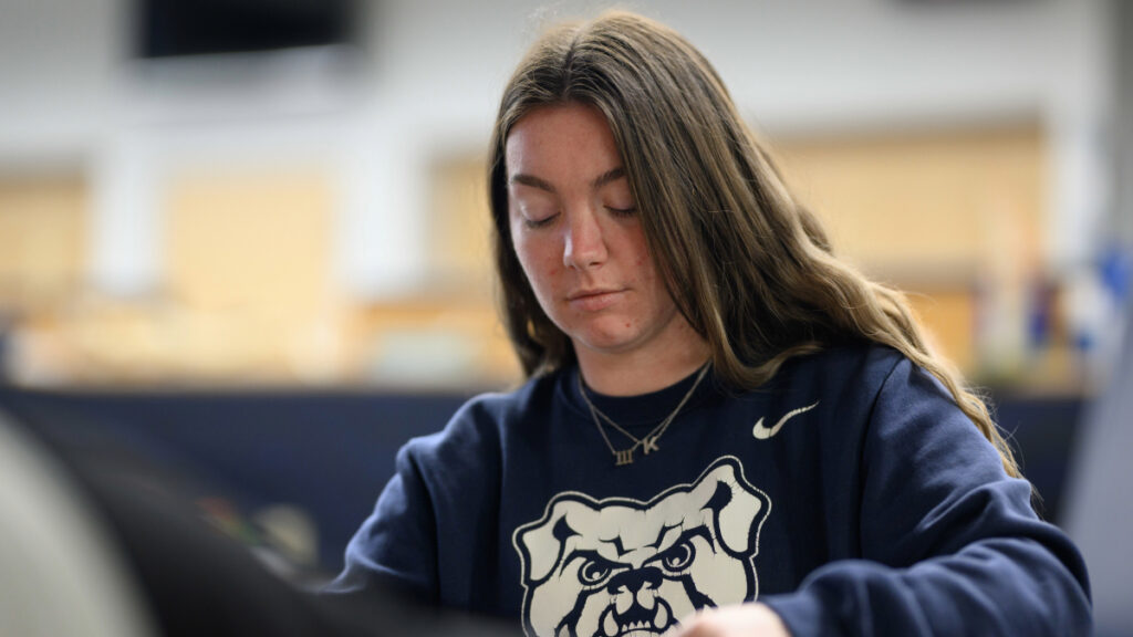 Female Butler student wearing a navy crewneck sweater with the Butler bulldog, sitting down and studying.
