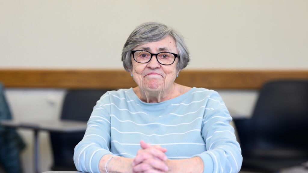 Nancy (McCoskey) Thoms ’75, MA ’24 wearing a light blue and white striped sweater, sitting in a classroom in Jordan Hall on Butler's campus.