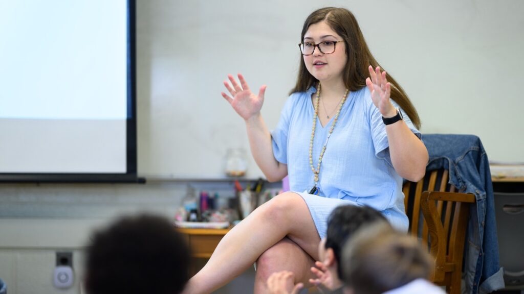 Butler student, Ashley Churchill '24, wearing a light blue short sleeved dress, and sitting on a wood chair teaching in an IPS/Butler Lab School #55 third grade classroom.