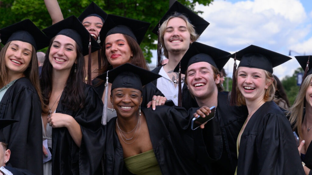 A group of eight Class of 2024 Butler graduates smiling and posing for a picture in their black caps and gowns.