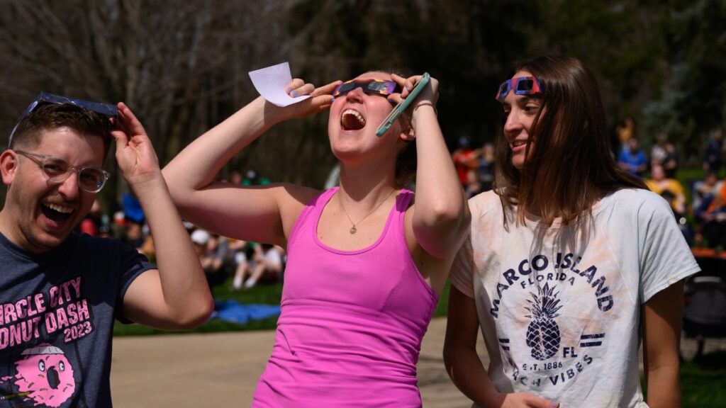A young male with glasses, standing with two young females; all three wearing their eclipse glasses, excited to experience the total solar eclipse from Butler's campus.