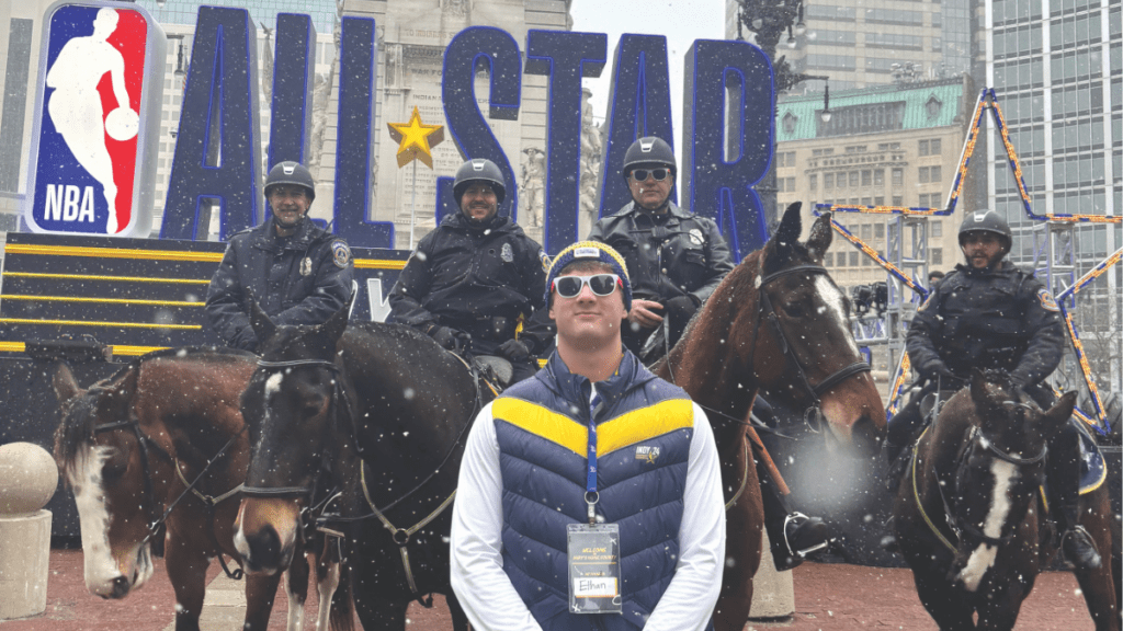 Butler student in a blue and yellow jacket standing in front of an NBA All-Star sign with four officers on horseback.