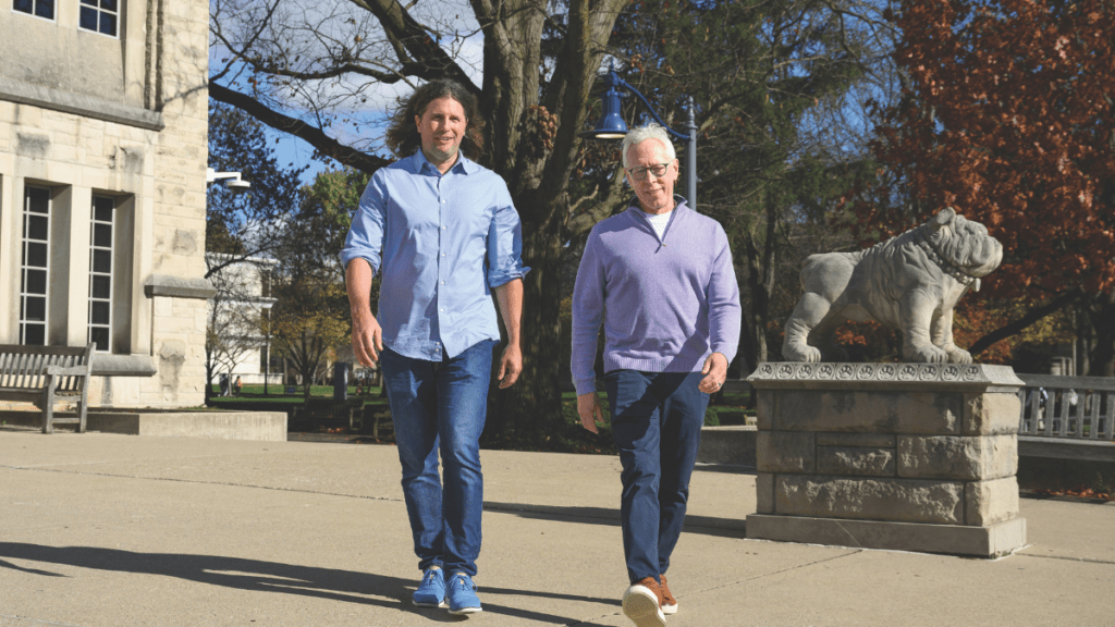 Randy Brown and Dr. Bryan Foltice walking near Atherton Union and the bulldog statue on Butler's campus, with autumn trees in the background.