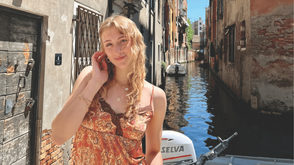 Caroline Erickson posing in a red and orange tank top, smiles by a canal lined with old buildings in Europe.