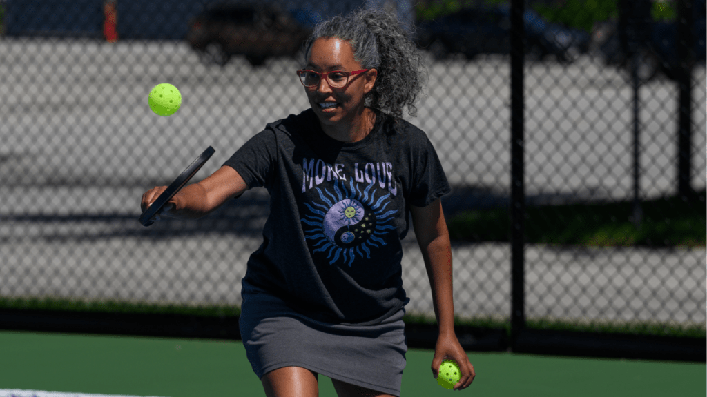 Person playing pickleball, holding a paddle and green ball, with a chain-link fence in the background.