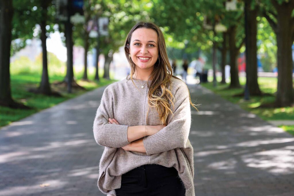 Kylie Stine smiles while crossing her arms with a shady tree-lined paved path behind her.
