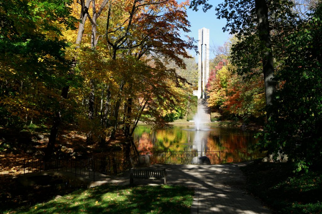 Autumn trees surround pond with active fountain and carillon behind it.