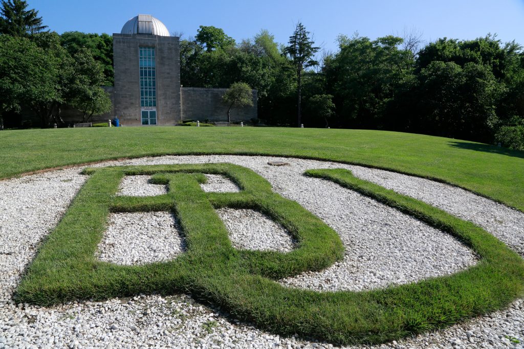 Large "BU" letters formed by green grass are outlined by white rocks on a hillside, with the Butler Observatory building behind it.