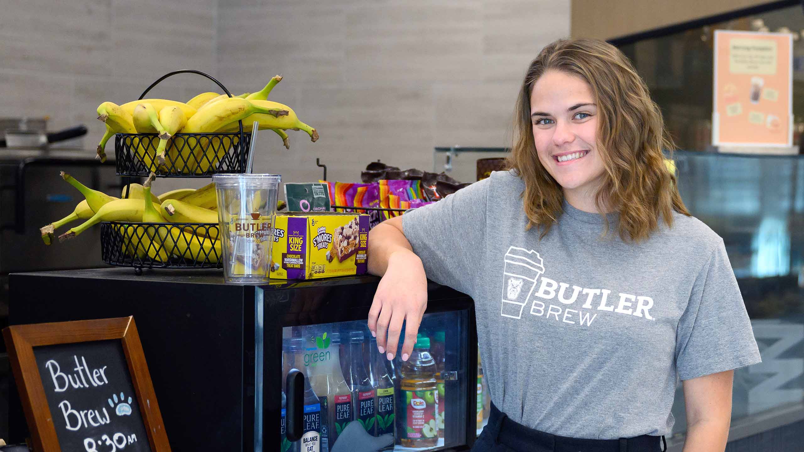 Female employee smiles while leaning on mini fridge at counter of Butler Brew coffee shop.