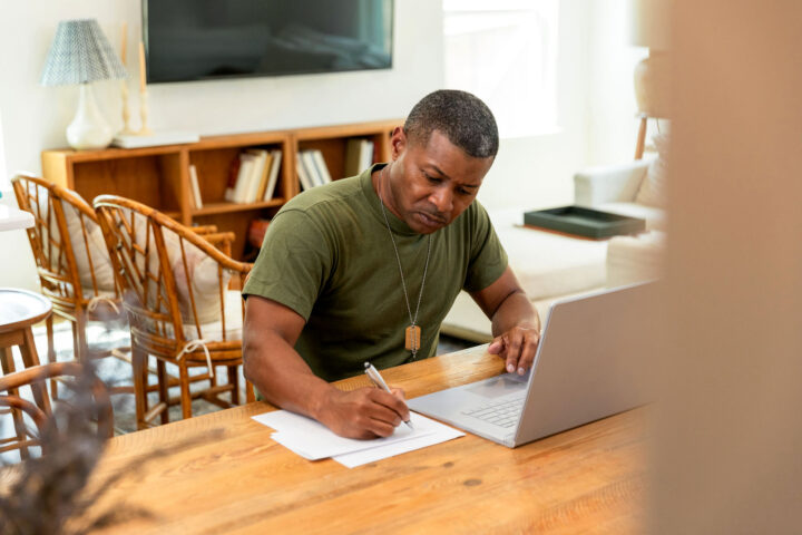 Male veteran writes notes while working on laptop at home.