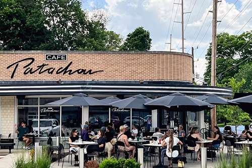 Groups of people sit at outdoor dining tables in front of a trendy cafe.