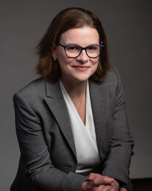 A formal headshot of a woman wearing a gray suit and blue glasses smirking at the camera. 
