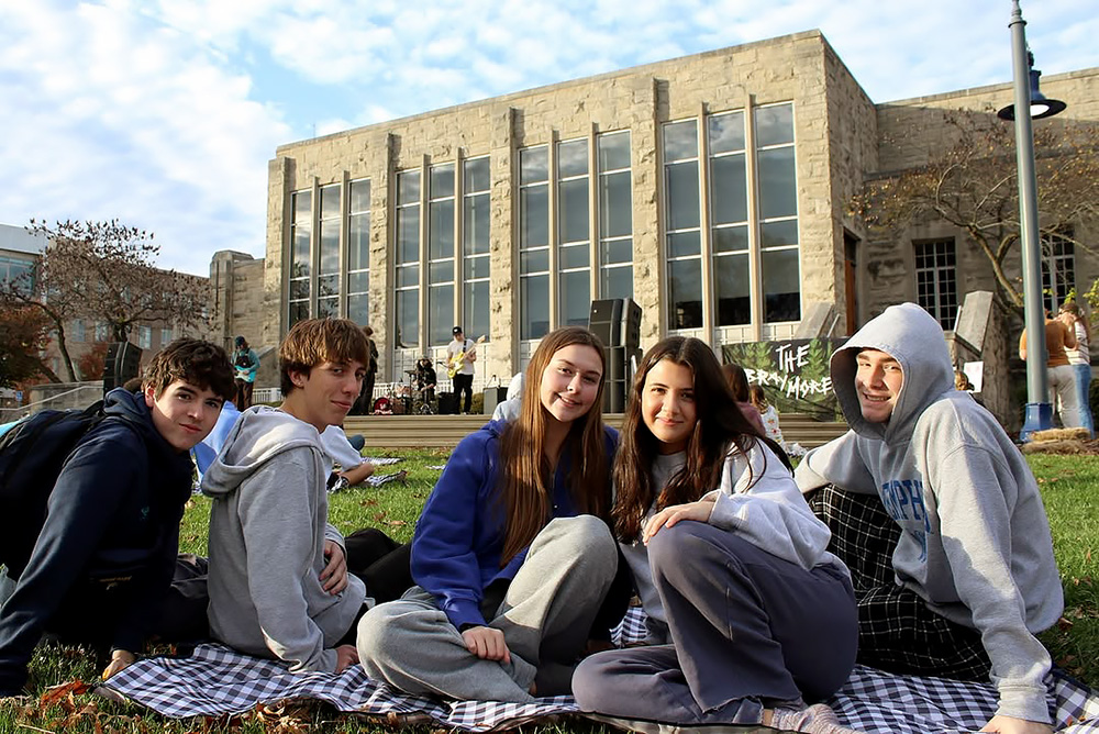Group of people smile for a photo while sitting on a picnic blanket at an open-air concert.