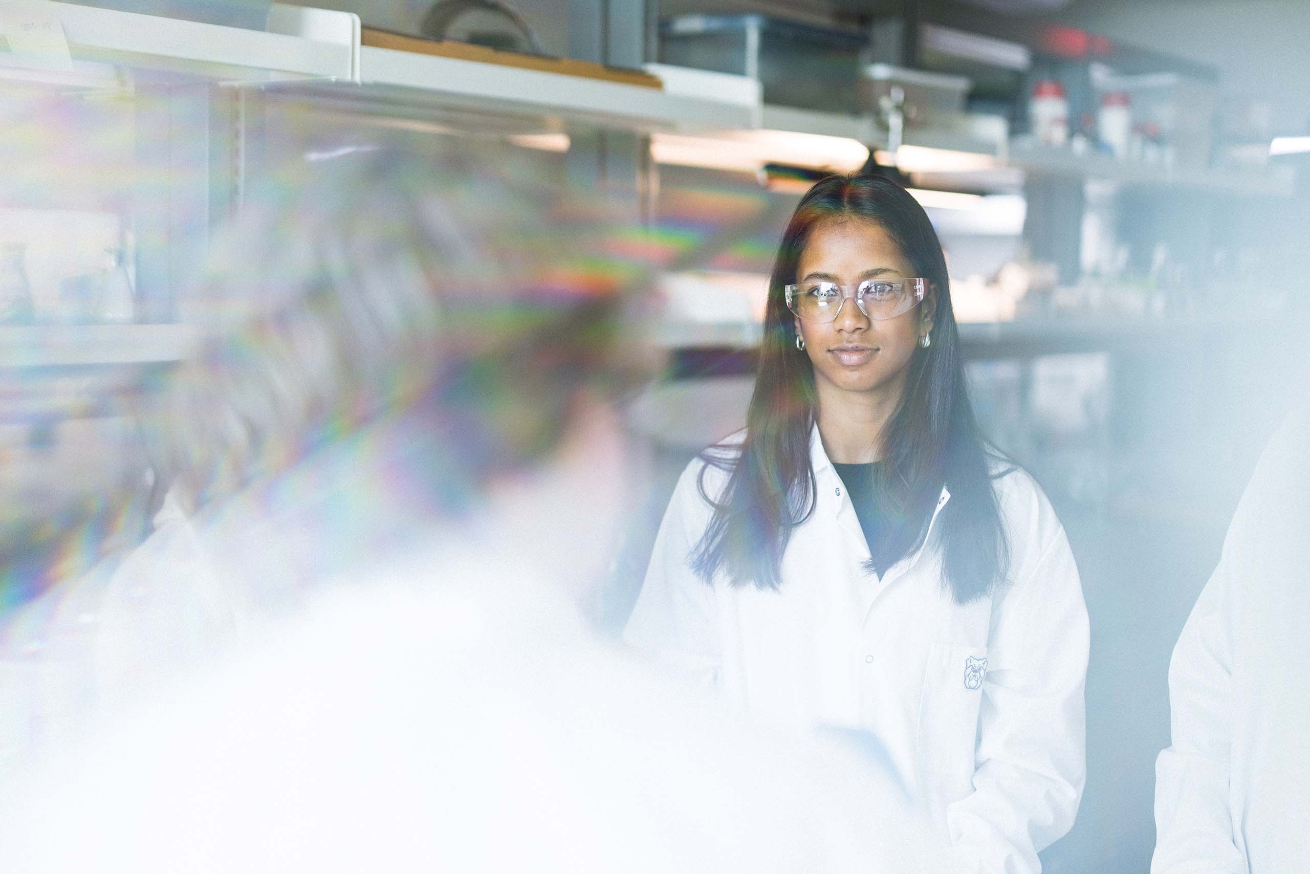Female college student in chemistry lab attire listening to professor.