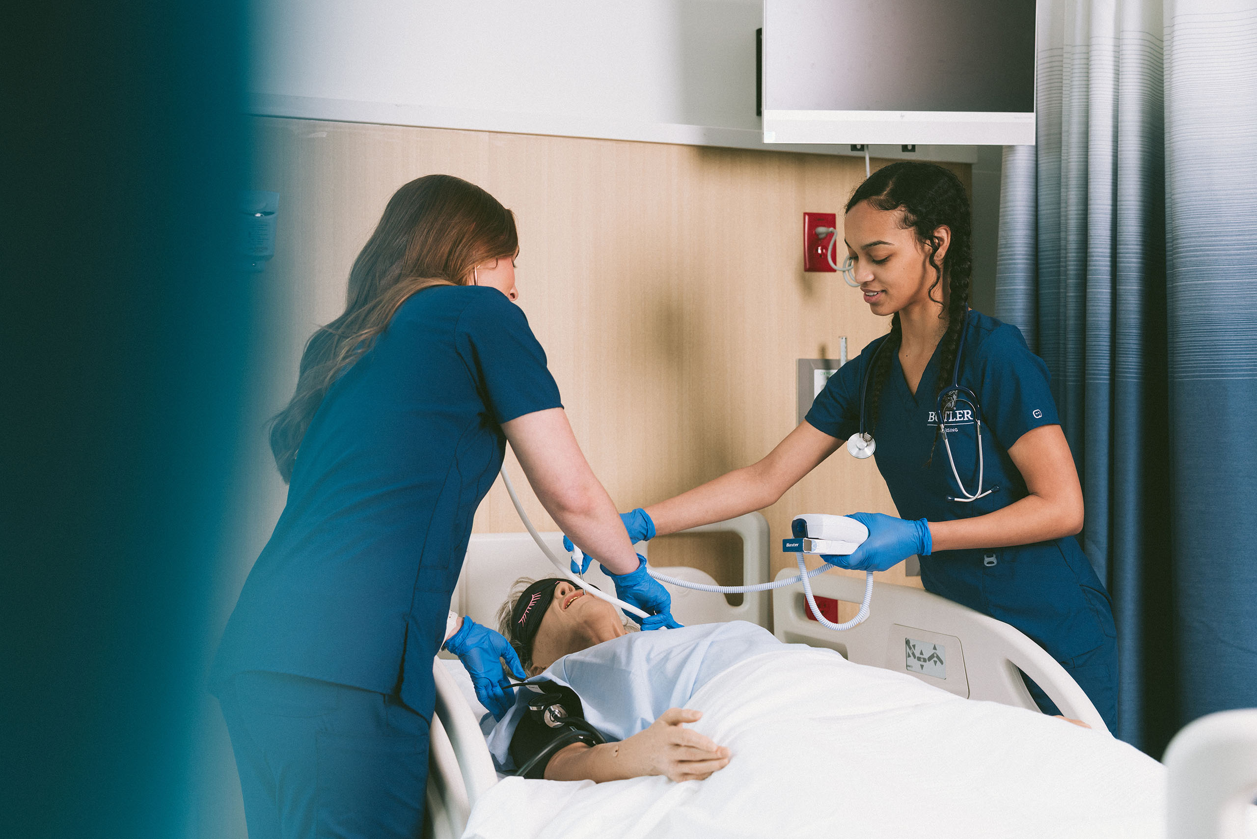 Two female nursing students tend to a patient lying down in a hospital bed.