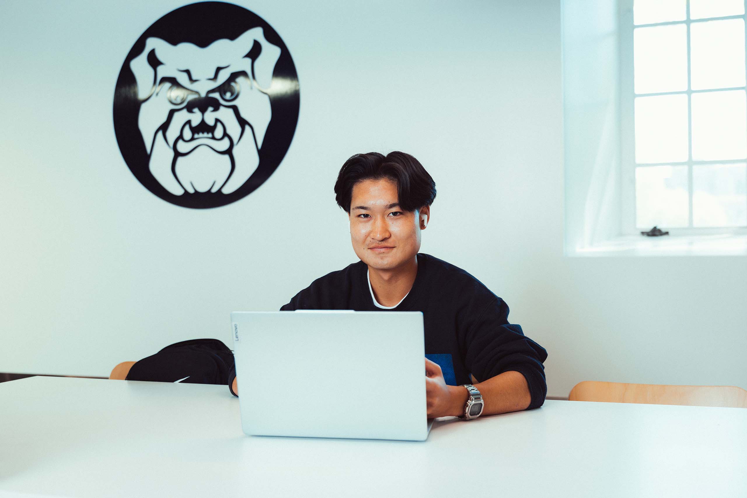 Portrait of male college student at laptop in a cafeteria with a Butler bulldog logo on the wall behind him.