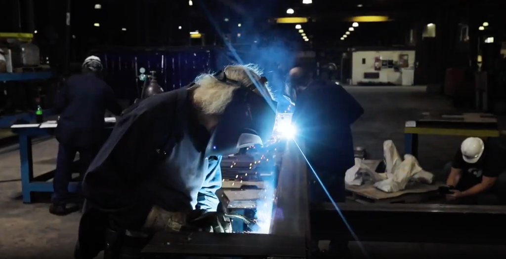 A masked metalworker causes sparks to fly while working on a project in a dimly lit workshop.