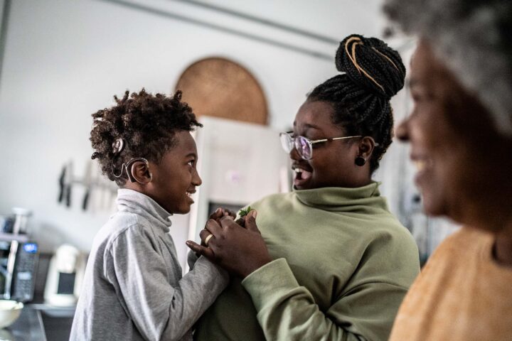 Mother and son with cochlear implant smile and hold hands at home with the grandmother smiling nearby.