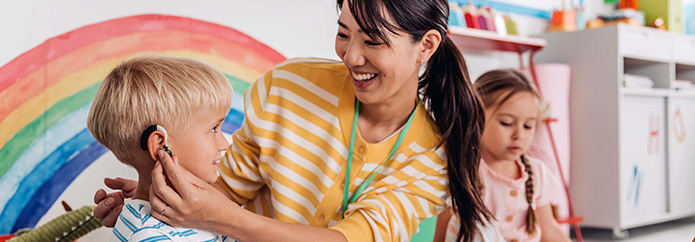 Teacher replaces hearing aid on young student while crouching on classroom floor.