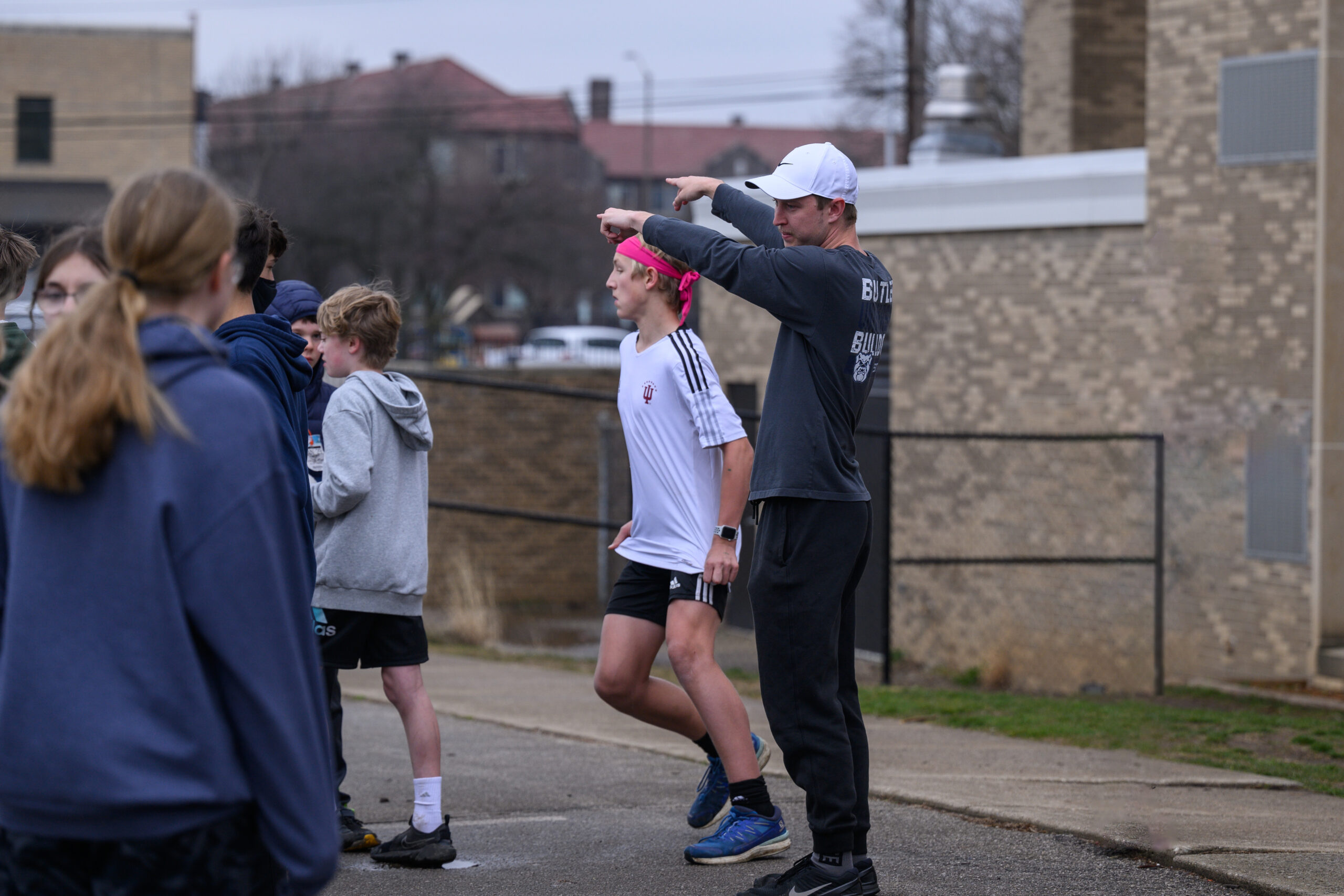 Male and female young students playing soccer, while college-aged man in blue shirt and white hat coaches them.