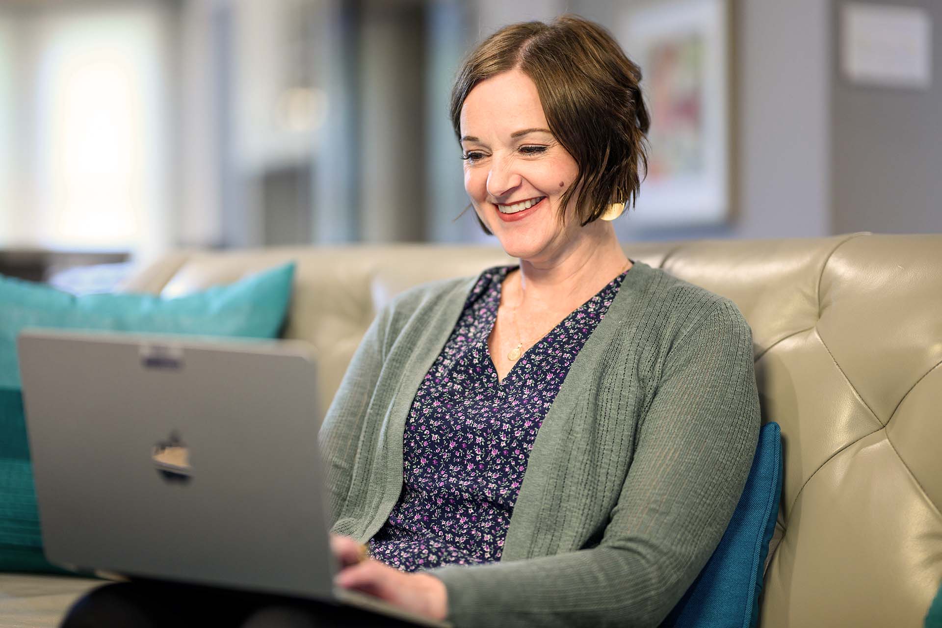 Smiling adult female student uses laptop for an online course at home on a sofa.