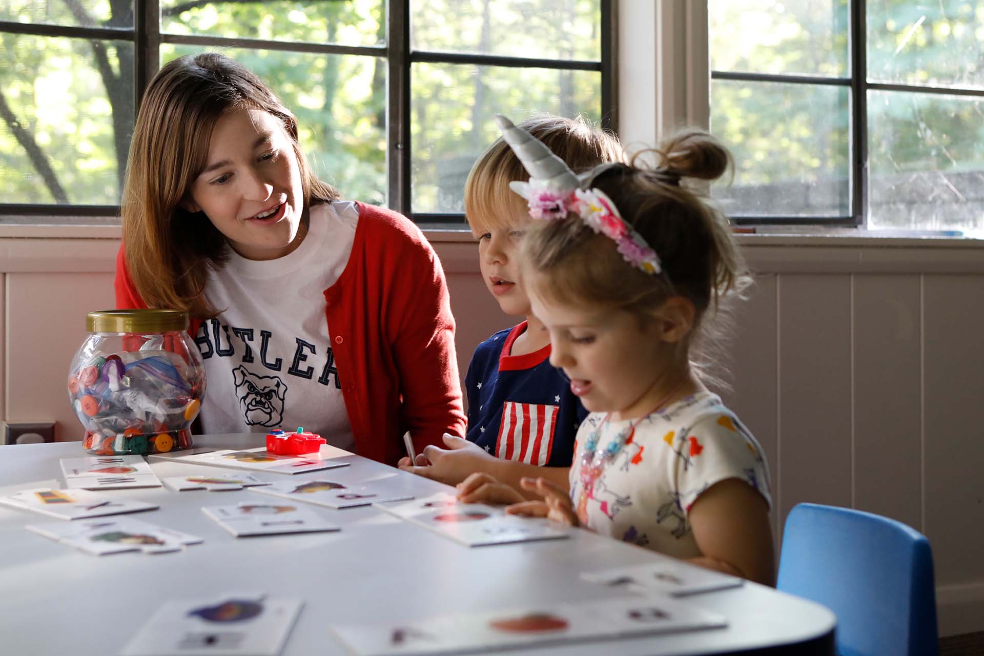 Teacher wearing Butler University shirt sits besides two young children at a table working on an activity.