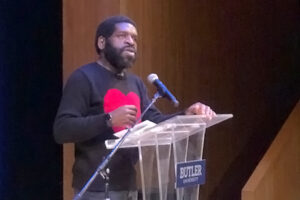 Hanif Abdurraqib speaking from plexiglass podium while wearing a sweater with a large red heart shape on the chest.