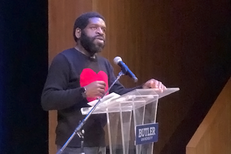 Hanif Abdurraqib speaking from plexiglass podium while wearing a sweater with a large red heart shape on the chest.
