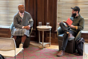 Rasheed Newson fields questions while sitting in a chair, listening attentively to man in the chair beside him.