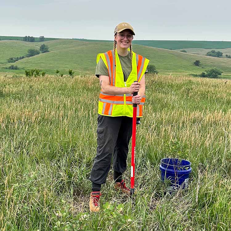 Natalie Long smiles while standing in lush, rolling hills with her archaeological technician vest, shovel, and bucket.
