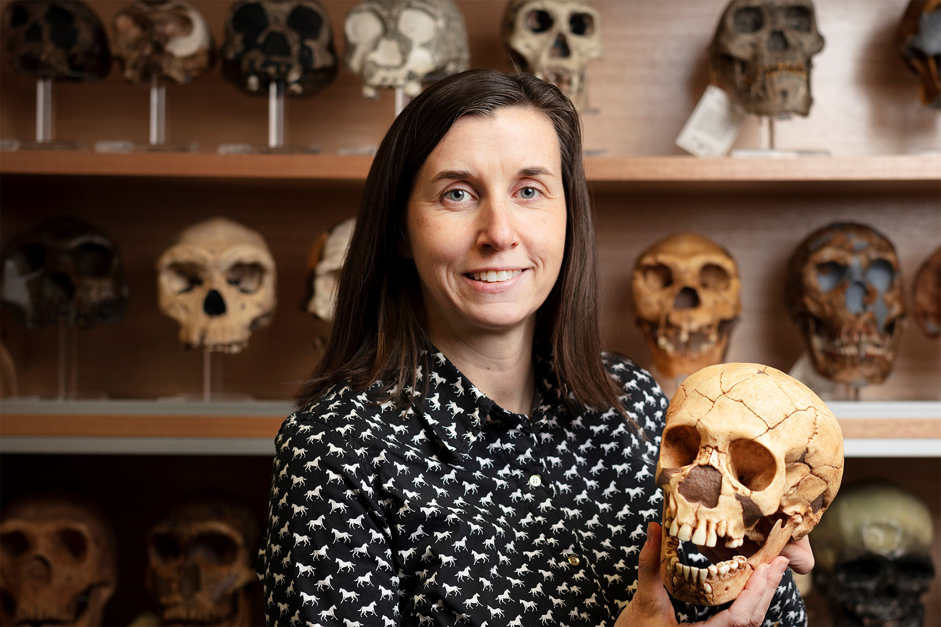 Woman holding a human skull in front of a shelf of many skulls.