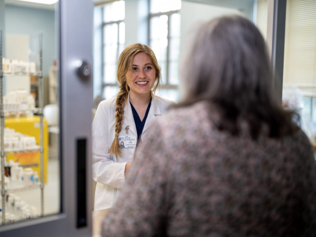 Smiling pharmacy student in white coat welcomes patient to pharmacy store room window