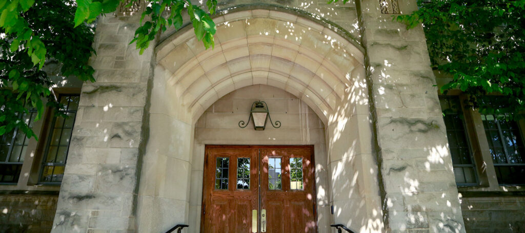 Pharmacy and Health Sciences Building arched entrance doorway with limestone walls and brown wooden doors is shaded by trees.