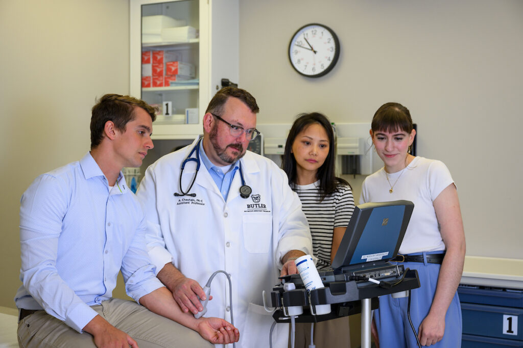 Three PA students, two young women and one young man, listen to a male PA preceptor in a white coat providing instruction in a clinical setting. All four professionals are gathered around a monitor on a medical device.