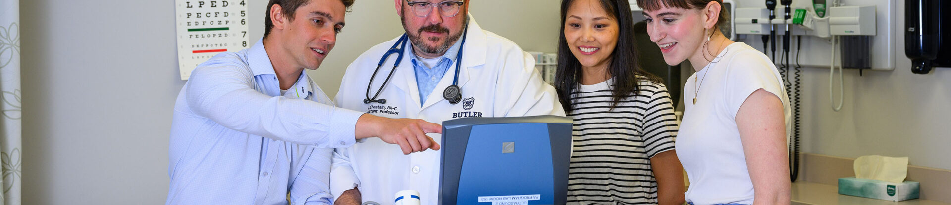 Multiple PA students look at monitor screen as professor uses device on a student acting as a patient in an exam room.