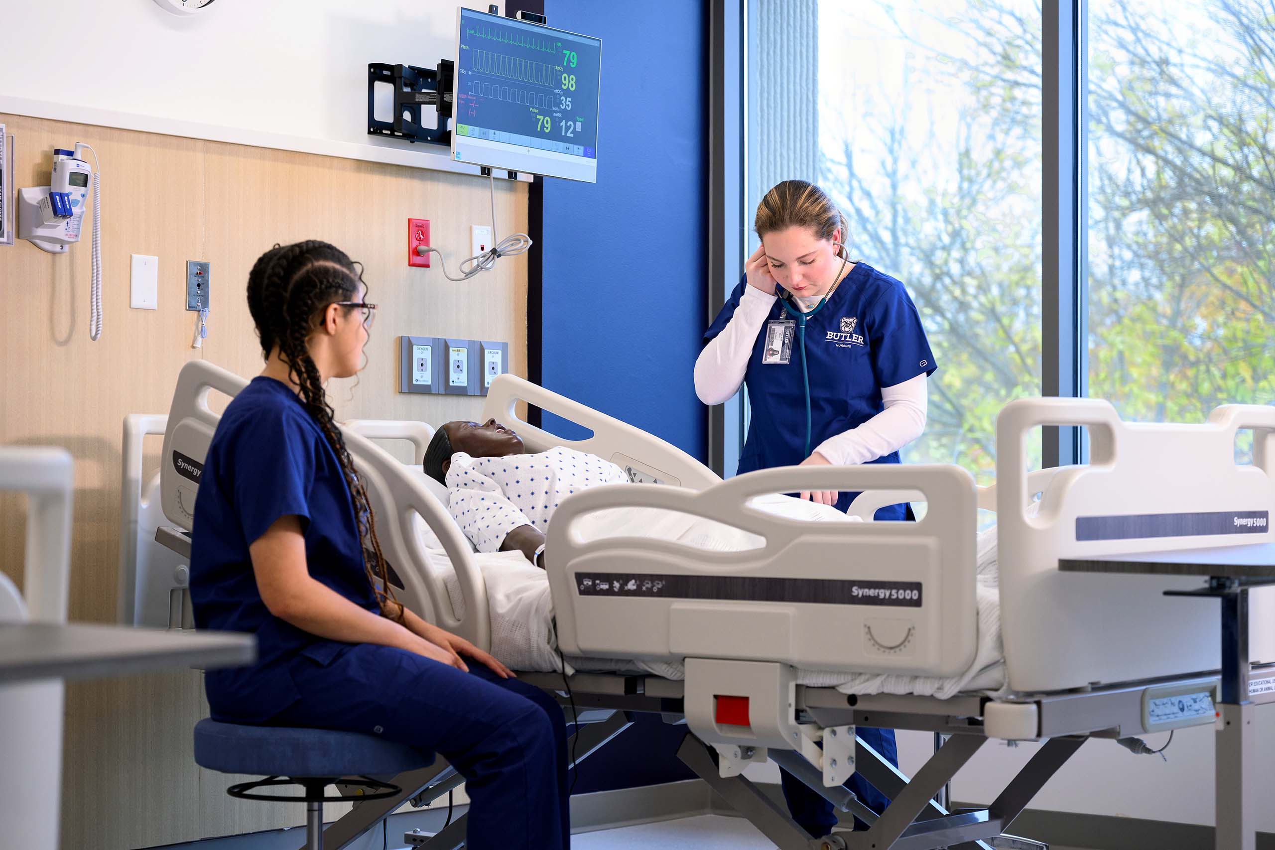 Nursing student listens to patient through a stethoscope at their bed while standing beside a vitals monitor screen.