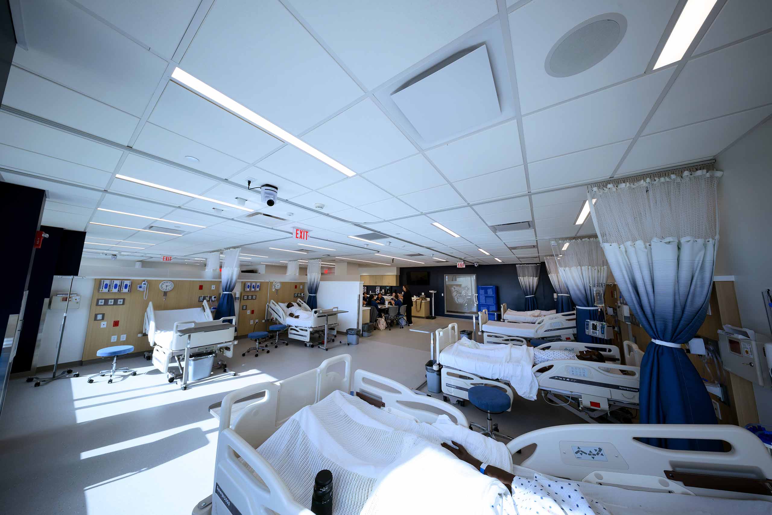 Wide view of mock hospital setup with rows of medical beds as nursing class meets at a table at end of room.