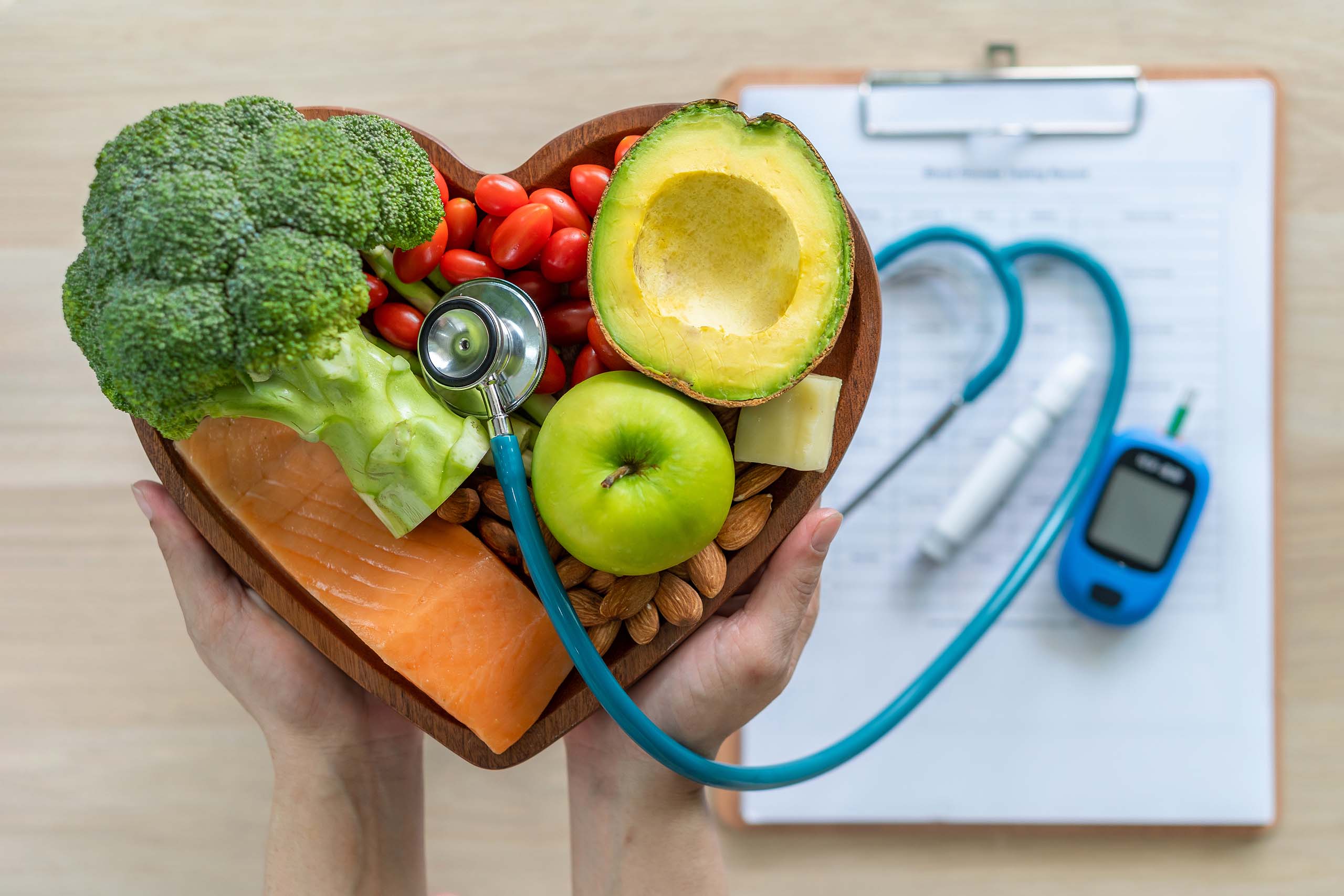 Healthy food in a heart-shaped bowl with medical equipment such as a stethoscope and patient paperwork on a clipboard.