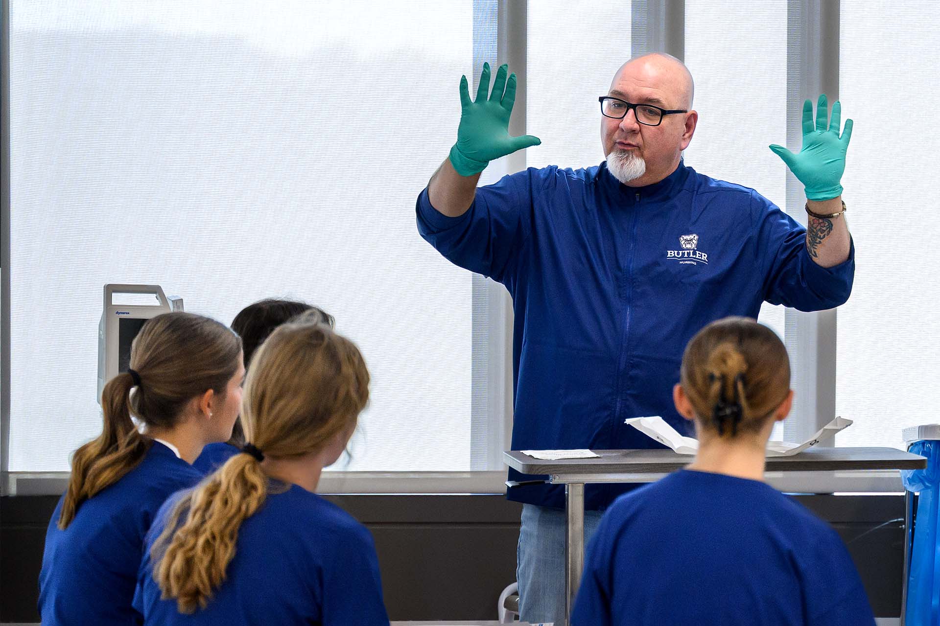 Nursing instructor wearing teal medical gloves shows his hands to a group of nursing students sitting around him.