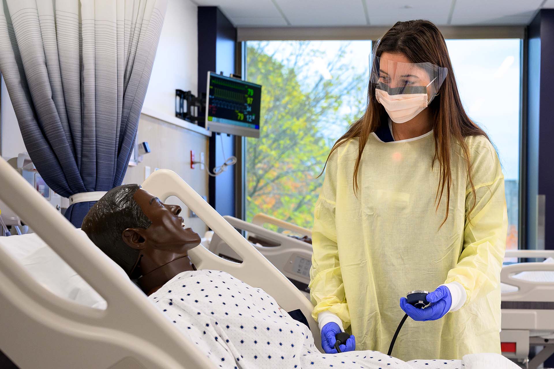 Nursing student in yellow gown and masks checks vitals of manikin patient in hospital bed.