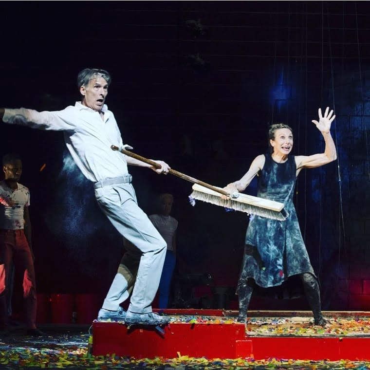 Male and female performs hold a large push broom on stage and have animated poses.