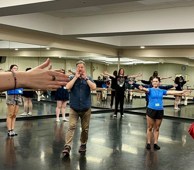 Instructor speaking to group of students with their hands extended in a practice room with mirrors on the walls.