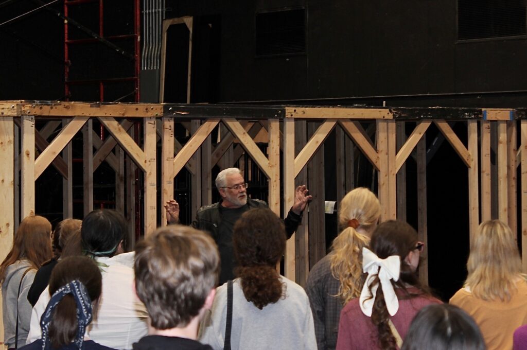 Instructor speaking to a group of students while in a backstage area with wooden frames behind him.